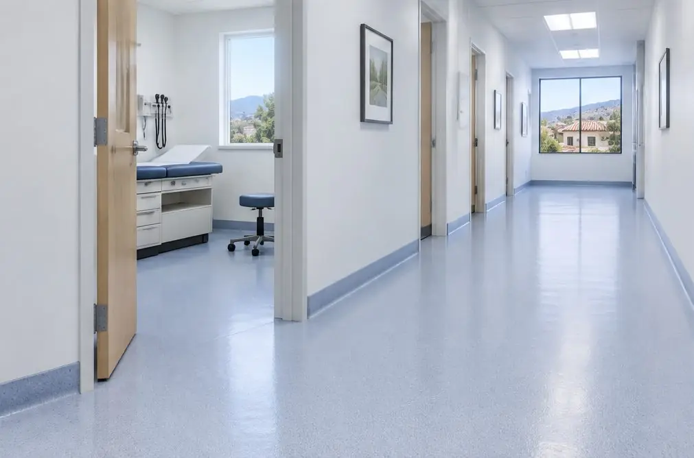 Illustration: a quiet clinic corridor with a seamless pale-blue-gray floor coating, a coved base at the wall and an open exam room door