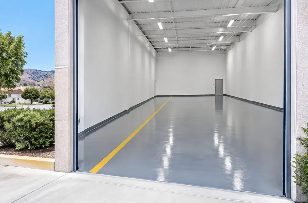 Illustration: a bright commercial interior floor in graphite epoxy with a painted yellow aisle line and a coved edge at the wall, seen from a loading dock door