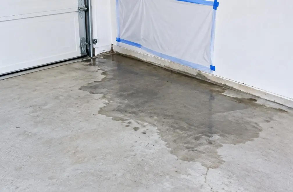 Illustration: a darker damp-looking patch spreading across a bare concrete garage slab near the wall, with a small drain grate nearby