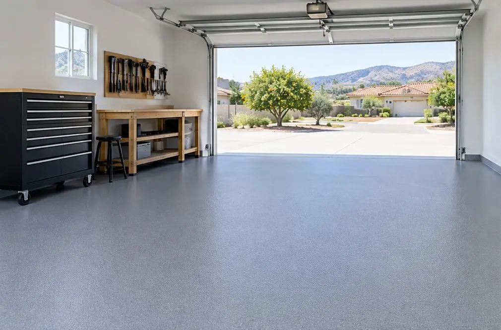 Illustration: a smooth mid-gray solid color epoxy floor in a narrow one-car garage with a workbench along one wall and the door open to daylight